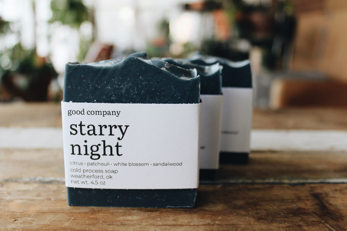 Three handmade soap bars wrapped in white paper labels reading 'good company starry night' sit on a wooden surface with a softly blurred indoor background.