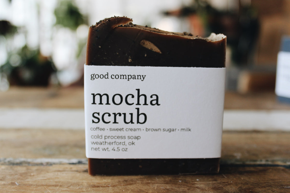 A bar of Good Company Mocha Scrub cold process soap standing upright on a wooden surface. The soap is dark brown with a white paper label that reads 'good company mocha scrub' and lists ingredients and location details.