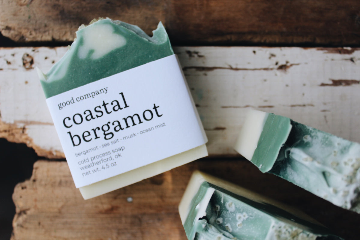 A bar of handmade soap labeled 'good company coastal bergamot' resting on a rustic white-painted wooden surface, with additional soap bars partially visible nearby.