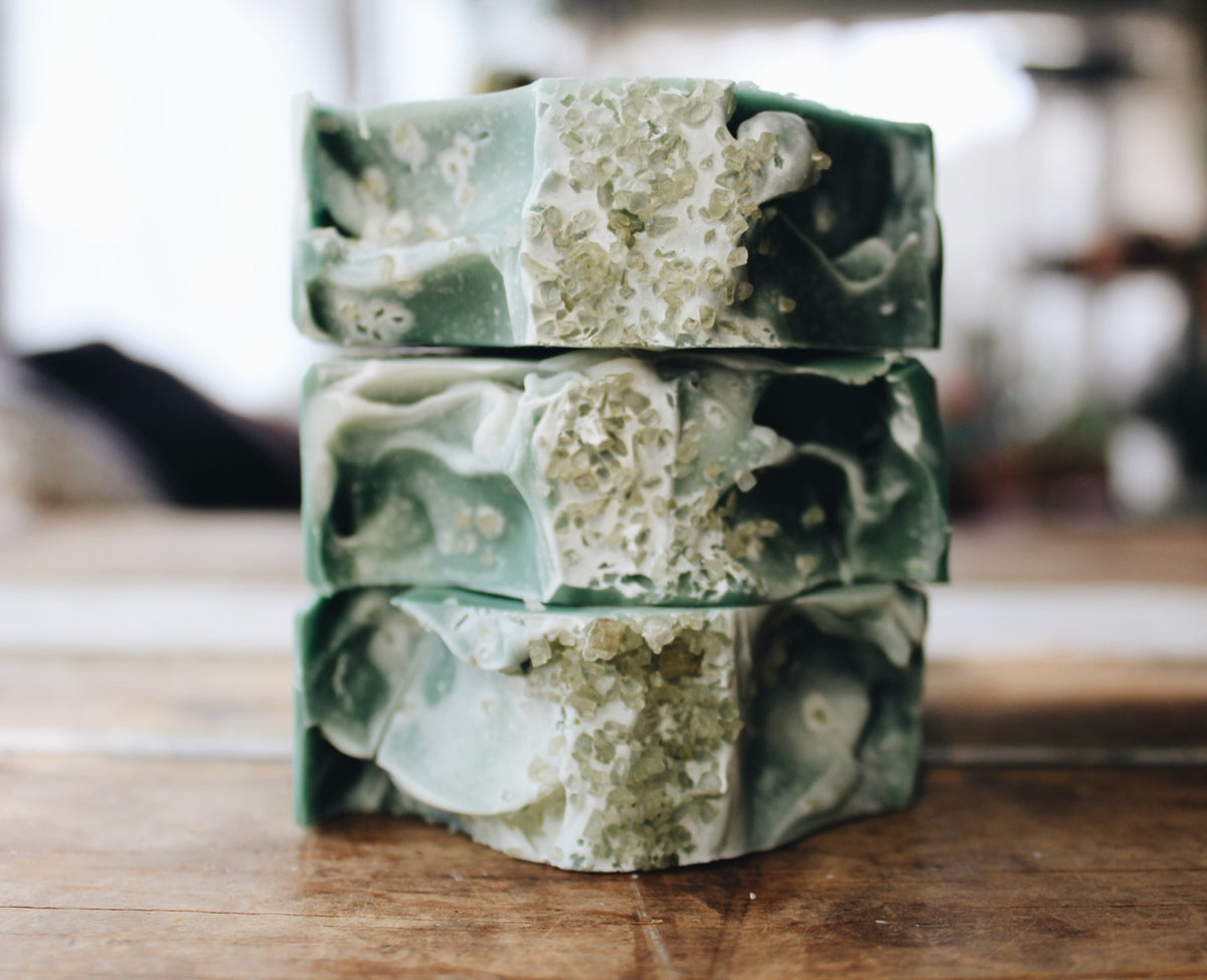 Three stacked handmade soap bars with a marbled green and cream design and embedded sea salt on top, set on a rustic wooden surface with a soft-focus background.