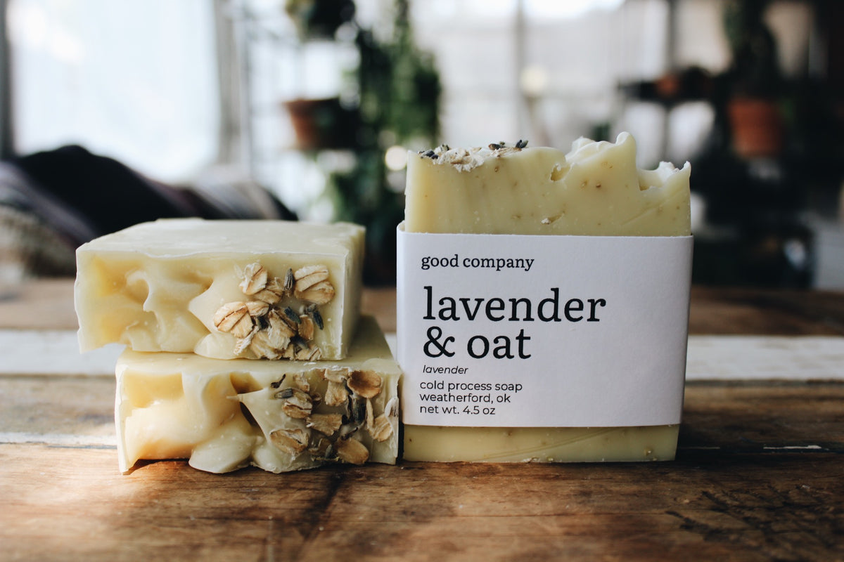 Three bars of handmade lavender and oat soap displayed on a rustic wooden table, with one bar in a white paper label on the right that reads 'good company lavender & oat'.