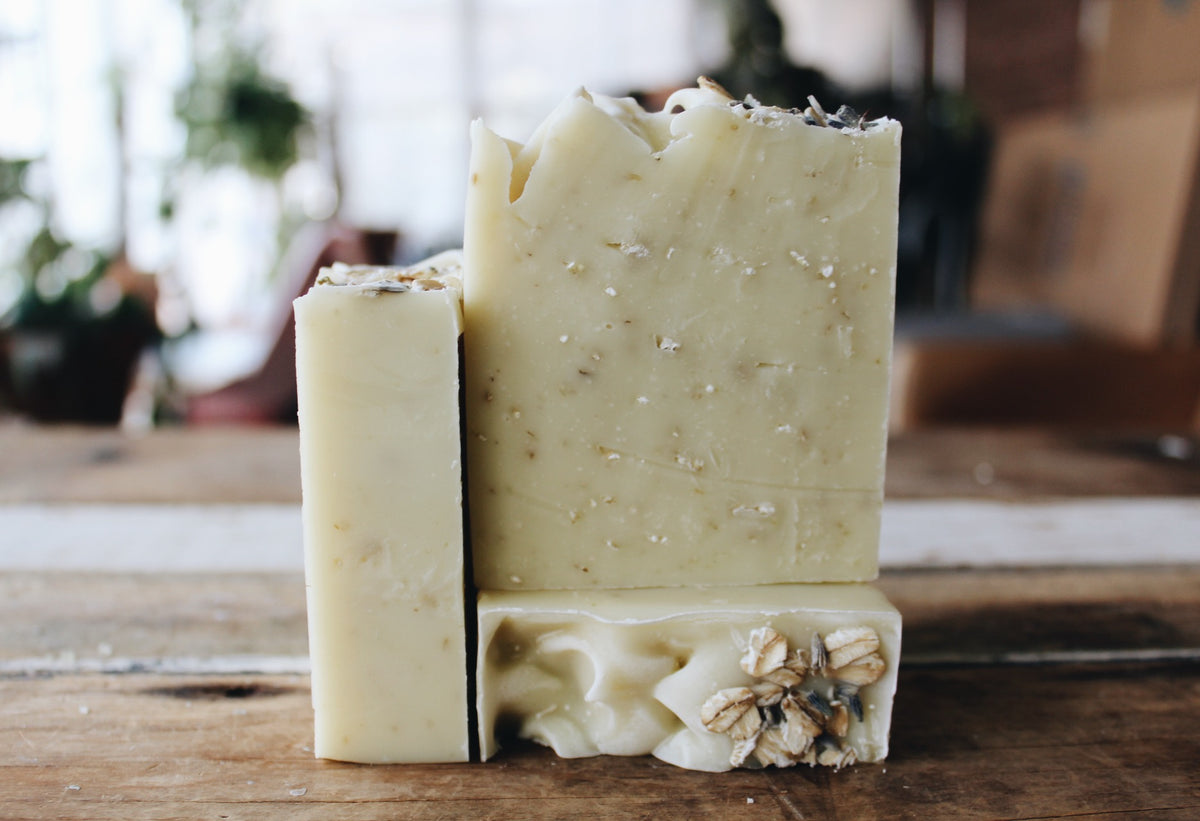 Three handmade soap bars with rough, rustic edges and embedded botanical/oil inclusions, standing on a wooden surface with a softly blurred indoor background.