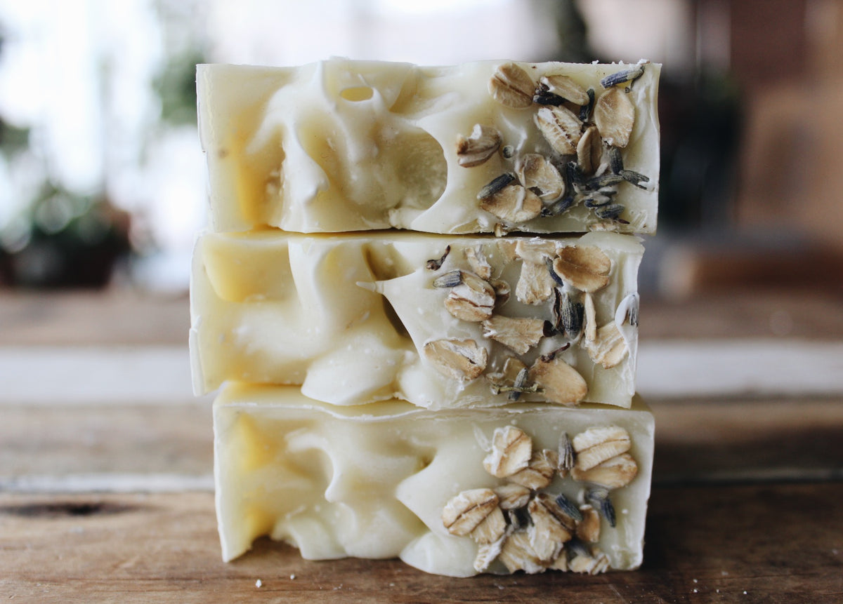 Three stacked handmade soap bars with a creamy off-white base and embedded oats and lavender buds, set on a rustic wooden surface with a blurred background.