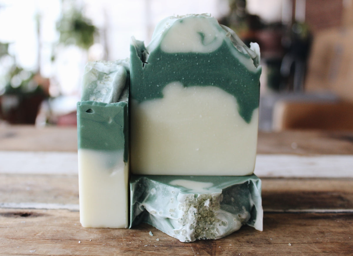 Three handmade soap bars with a green and cream marbled swirl pattern, displayed on a rustic wooden surface with a blurred indoor background.