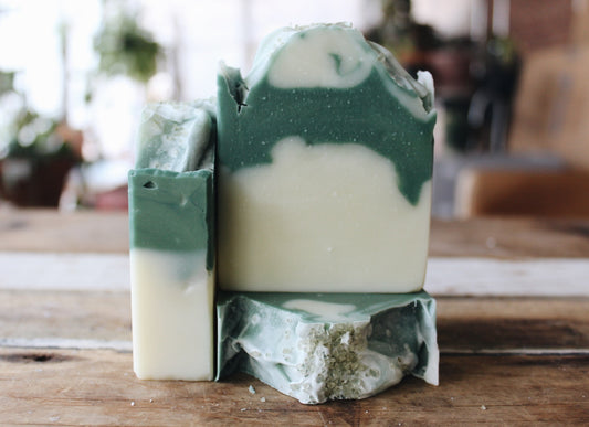 Three handmade soap bars with a green and cream marbled swirl pattern, displayed on a rustic wooden surface with a blurred indoor background.