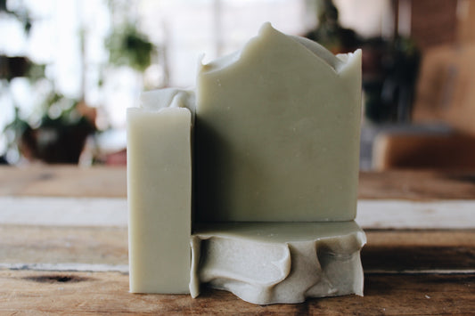 Three handmade soap bars made with clay in natural light that are muted sage green in color arranged on a rustic wooden surface with a softly blurred indoor background.