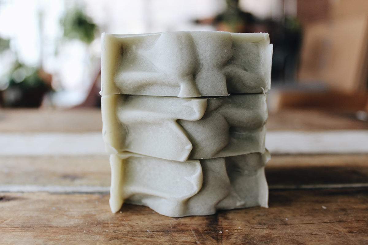 Three stacked handmade soap bars with a textured, organic top in a muted sage green color, set on a rustic wooden table with a soft-focus background.
