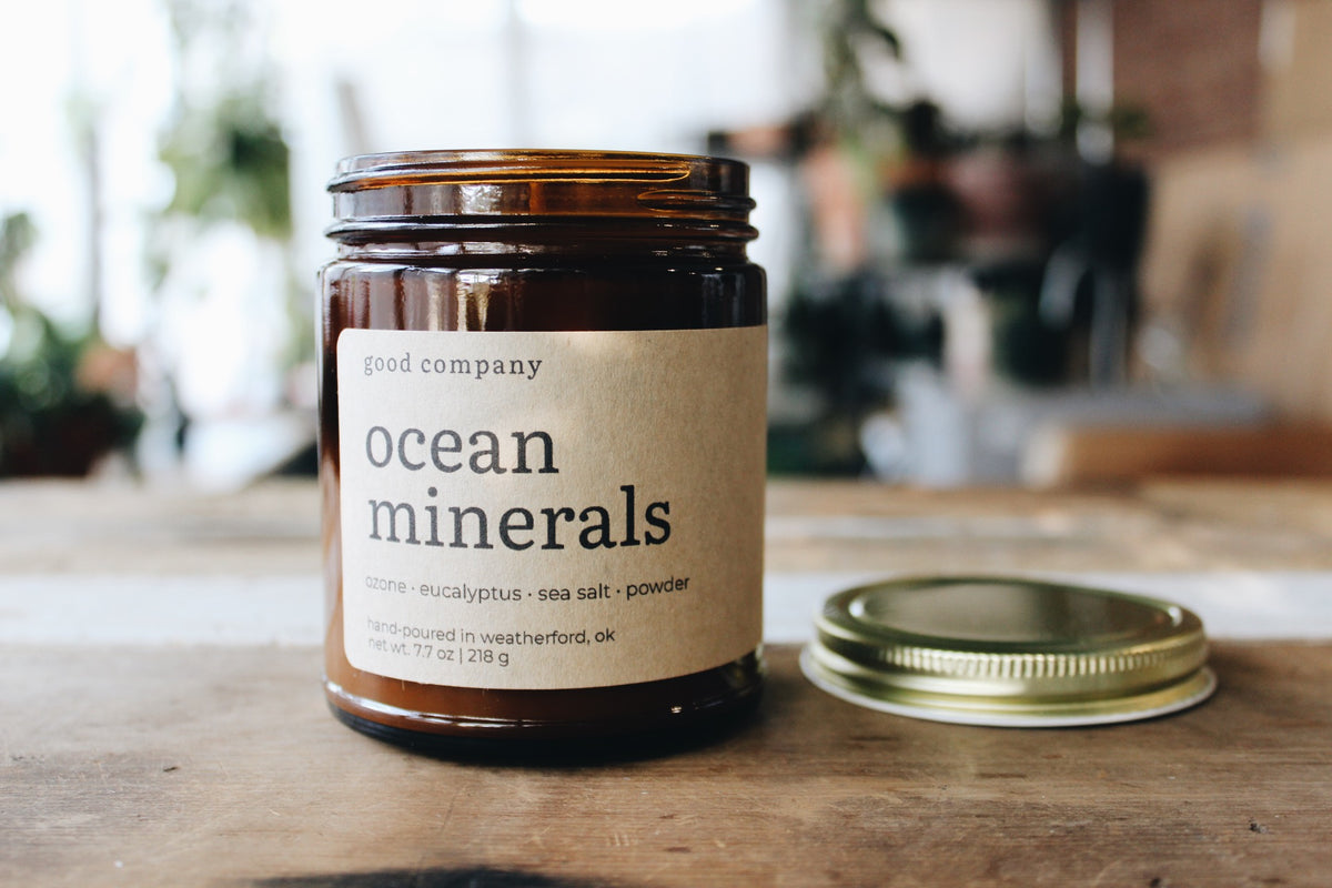 An amber glass jar labeled 'good company ocean minerals' sits on a wooden table with its gold metal lid placed beside it. The background is softly blurred, showing an indoor setting with plants and warm lighting.
