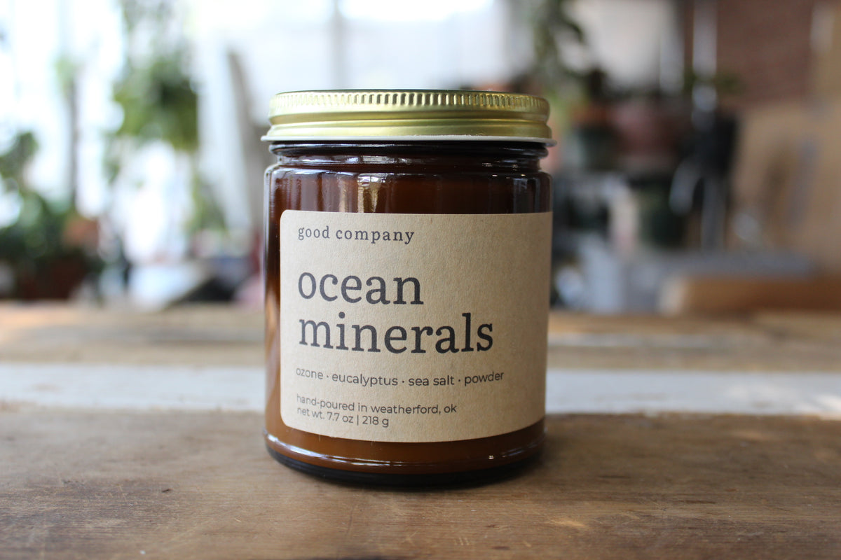 A glass jar of 'good company' Ocean Minerals candle featuring a kraft paper label and a gold metal lid, set on a wooden table with a blurred indoor background.